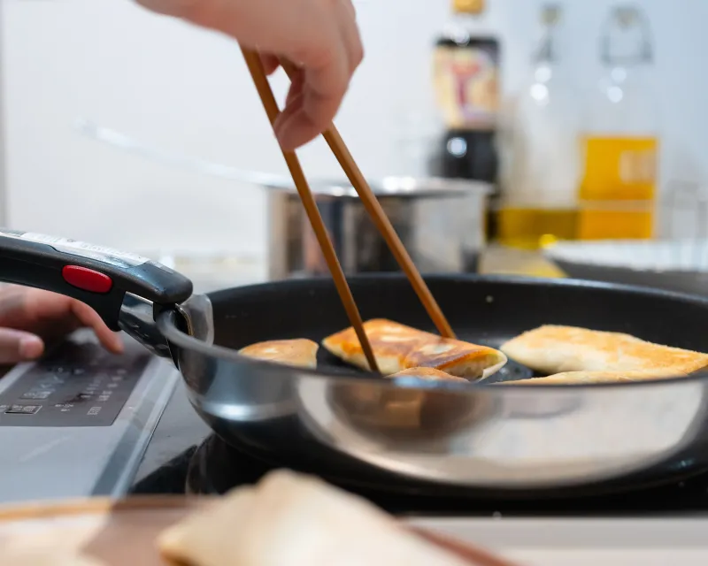 A hand using wooden chopsticks to flip golden pan-fried pancakes in a skillet on a kitchen stove