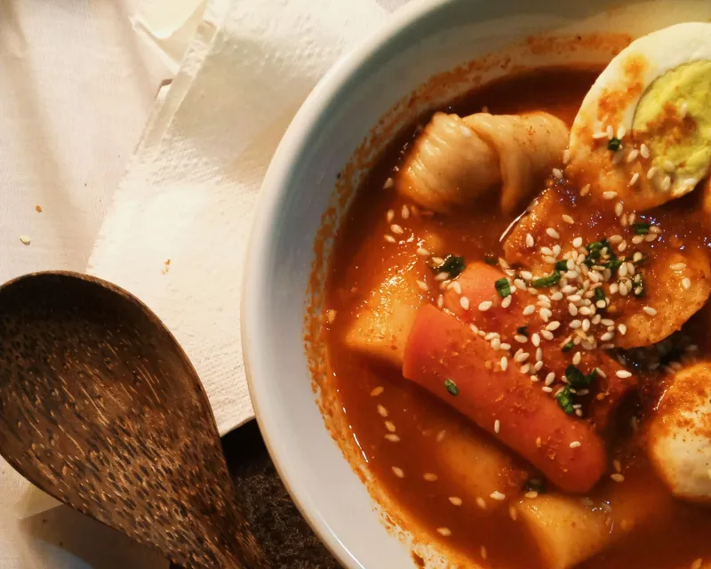 A bowl of tteokbokki rice cakes, fish cake and half a boiled egg in red chili sauce, with a wooden spoon beside it