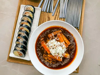Bowl of tteokbokki in dark chili sauce topped with white shreds, beside a tray of sliced gimbap and chopsticks