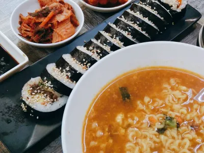 Sliced gimbap rolls on a black tray beside a bowl of noodle soup, with kimchi at the top