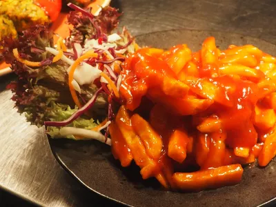 Glossy red tteokbokki rice cakes on a dark plate beside a small salad