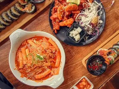 Overhead view of a wooden table set with a bowl of tteokbokki, a plate of fried chicken, gimbap rolls and small side dishes