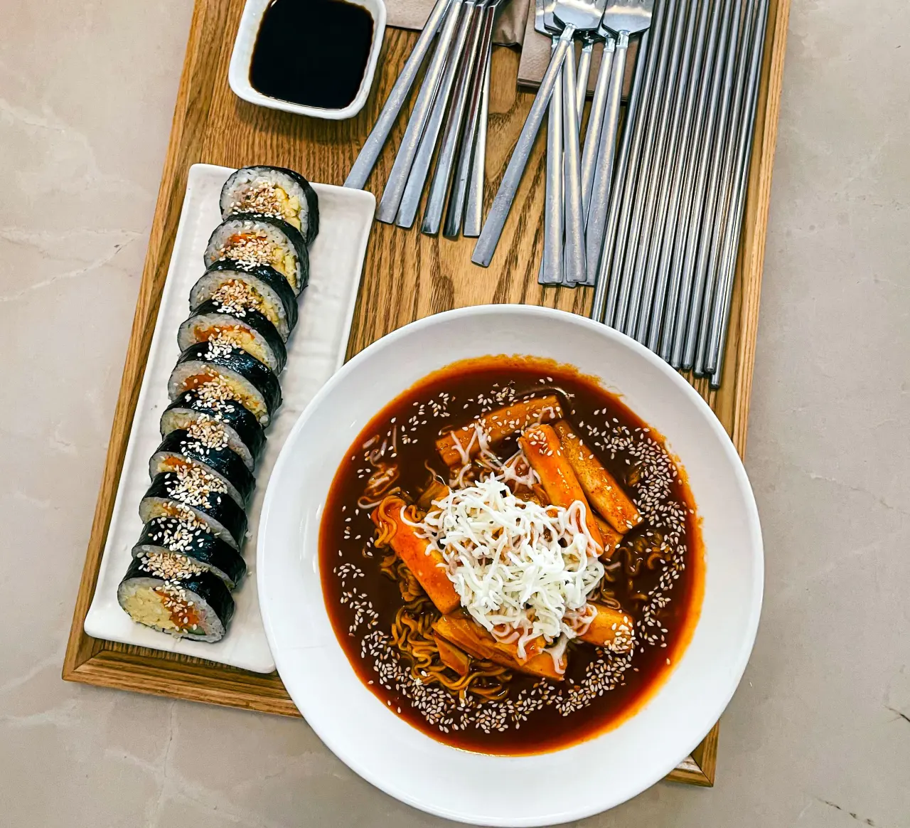 Overhead view of a wooden tray with sliced gimbap, a bowl of tteokbokki in dark chili sauce, soy sauce and metal cutlery