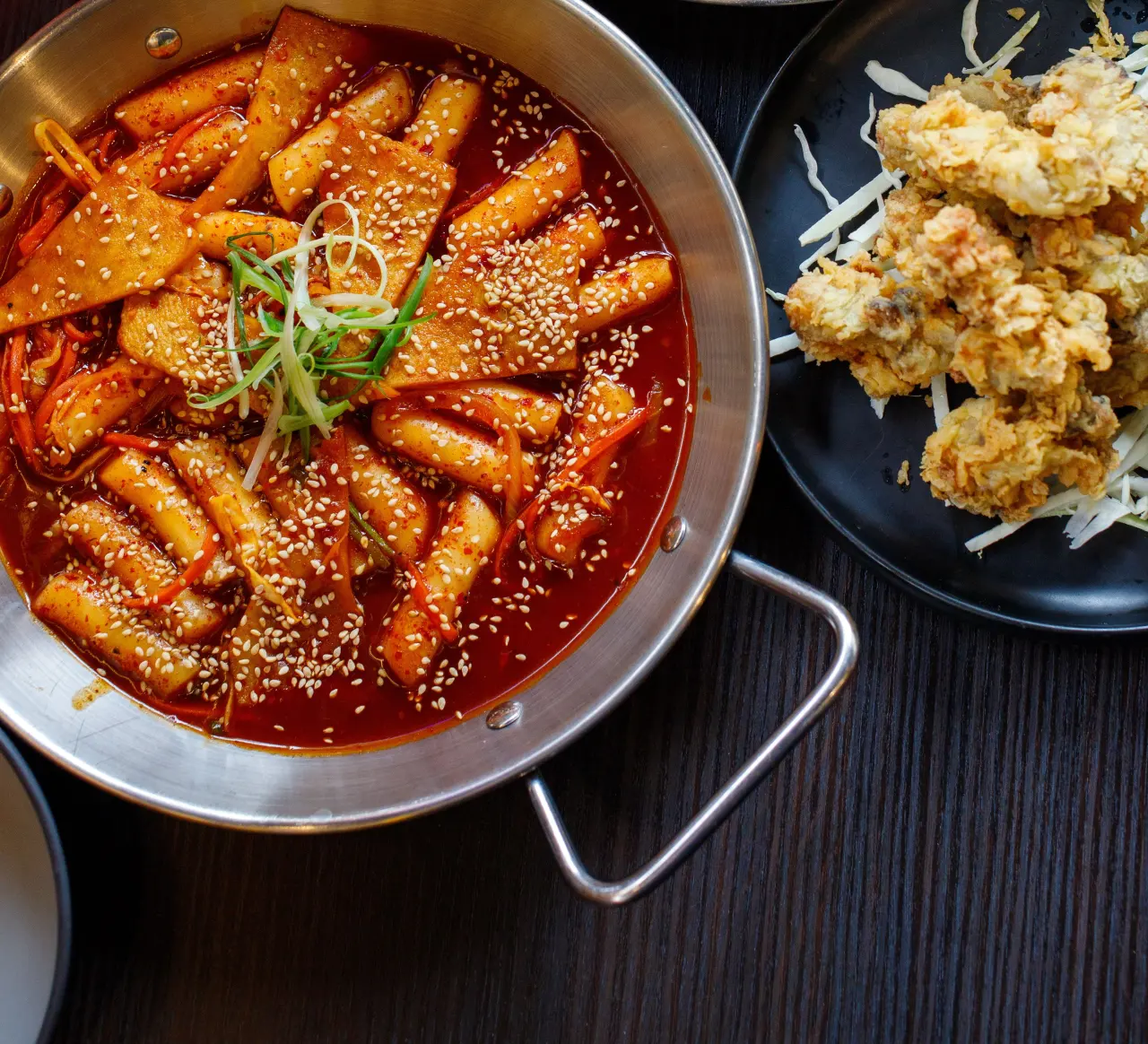 Overhead view of a steel pan of spicy tteokbokki with sesame seeds, beside a plate of fried chicken on a dark table